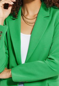 Zielona marynarka z wyraźnymi ramionami i gładką teksturą. Trzy warstwowe złote naszyjniki oraz biały top pod spodem.