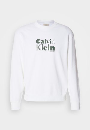 Bela trenerka z rebrastimi manšetami in spodnjim robom, ki na sprednji strani vsebuje velik zeleni napis "Calvin Klein" v moderni pisavi.