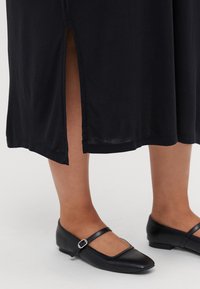 Musta, nilkkapitkä mekko, jossa on sivuhalkio ja pehmeä rakenne. Yhdistetty mustiin, tekonahkaisiin tasapohjaisiin kenkiin, joissa on neliön muotoinen kärki ja solkidetali.