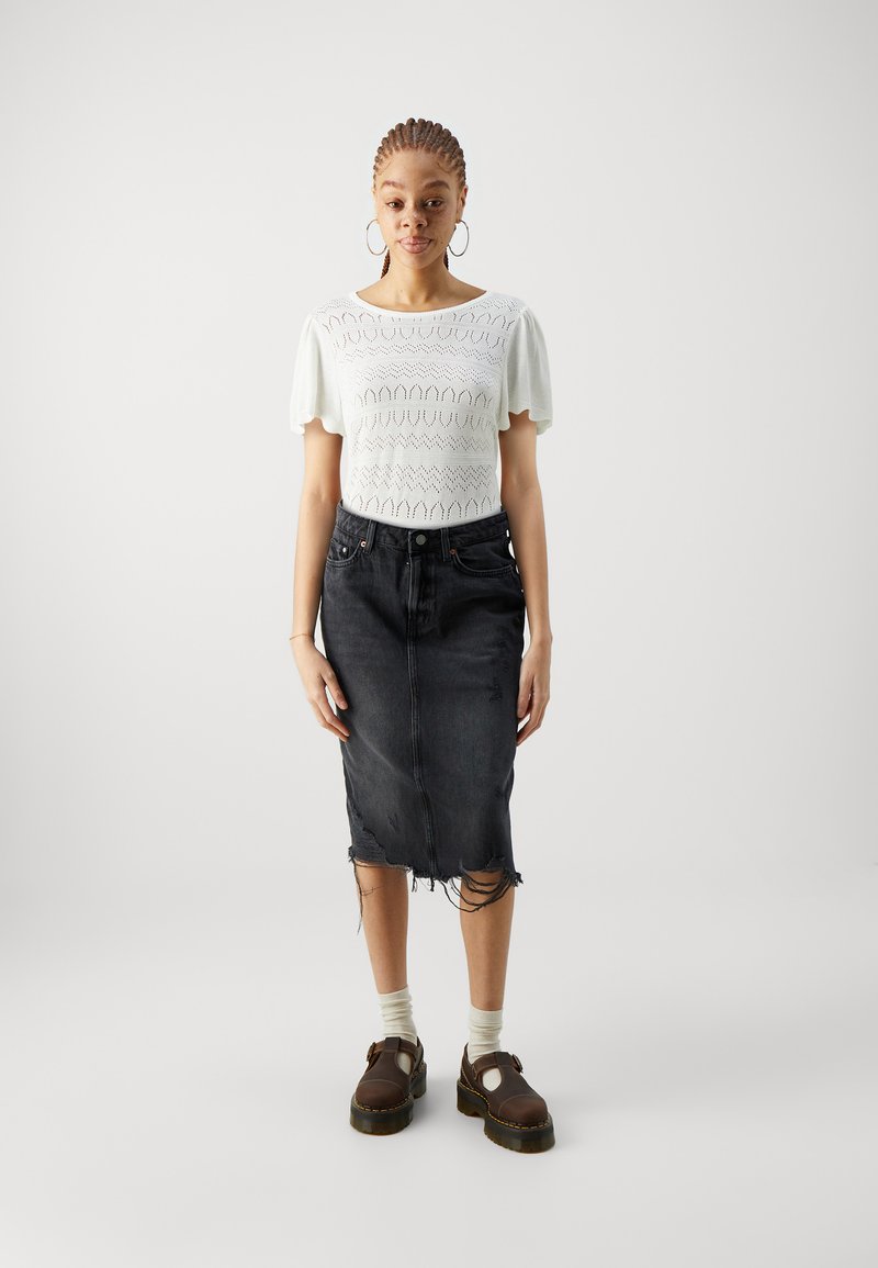Witte gebreide top met pofmouwen, gecombineerd met een zwarte denim rok met versleten rand en beschadigde details, en bruine platform schoenen.