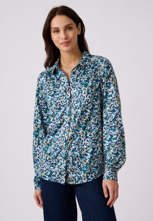 Femme aux longs cheveux foncés portant une chemise à manches longues bleu, blanc et orange à motifs avec des boutons et un pantalon bleu foncé.