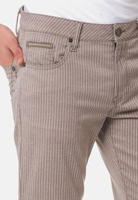 Hellbraune gestreifte Hose aus weicher Stoffqualität, mit einer Vordertasche, subtilen Steppdetails und einem Metallknopfverschluss.