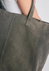Olivgrön mocka tote-väska med vertikal ribbad textur, en ensam axelrem och platt botten. Har en minimalistisk design och prydlig sömnad.