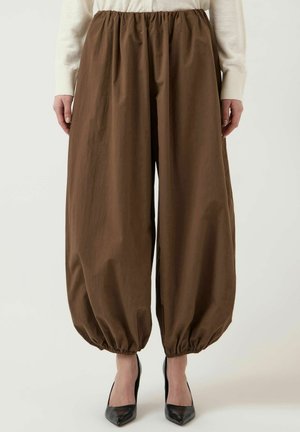 Kobieta ubrana w luźne brązowe spodnie ze ściągaczem u dołu, białą bluzkę z długim rękawem oraz czarne szpilki z zaostrzonym noskiem, stojąca na jednolitym tle.