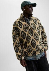 Fleece-Pullover in Beige mit schwarzen Rautenmustern. Hoher Kragen, halber Reißverschluss und weiche Textur. Gewählt über einem Denim-Hemd und dunklen Hosen.