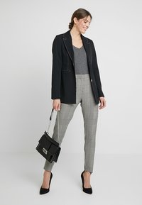 Juodas švarkas su baltais apvadais, pilkas topas ir languotos kelnės. Aksesuarai apima juodą rankinę su studijų akcentais ir juodus kulnus.