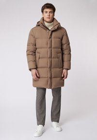 Giacca a vento imbottita di colore marrone chiaro, con cappuccio, chiusura zip, grandi tasche e design trapuntato. Abbinata a pantaloni grigi e sneakers bianche.