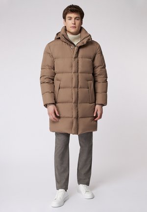 Giacca a vento imbottita di colore marrone chiaro, con cappuccio, chiusura zip, grandi tasche e design trapuntato. Abbinata a pantaloni grigi e sneakers bianche.