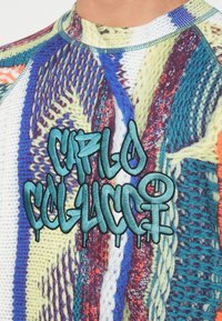 Mønstret sweater i flere farver med et struktureret strikkemønster og blå graffiti-lignende broderi, der står "CAYLO COLUCCI." Ribbestrikket krave og ærmer.