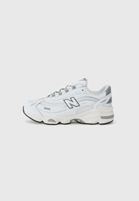 Chaussure de sport blanche avec des superpositions en mesh et en matériau synthétique, de forme courbée, avec des accents gris, une semelle épaisse et un logo "N" bien visible sur le côté.