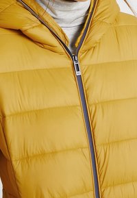 Chaqueta de plumas amarilla con capucha, con cremallera frontal en color azul marino, textura acolchada con costuras horizontales y un acabado suave.