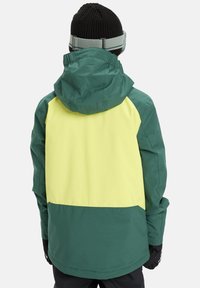 Kurtka w kolorach zielonym i żółtym w stylu blokowym, wyposażona w odpinany kaptur, elastyczne mankiety oraz kontrastujące czarne rękawiczki. Teksturowana tkanina.