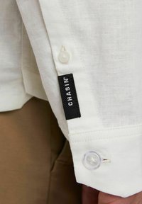Polsino di camicia in lino bianco con un bottone rotondo trasparente e un'etichetta nera cucita sopra, con il testo "CHASIN'." La texture è visibile.