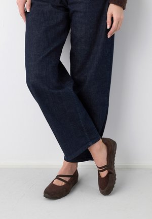 Personne portant un jean large et sombre et des chaussures fermées marron avec des brides croisées, debout sur un sol clair contre un mur blanc.