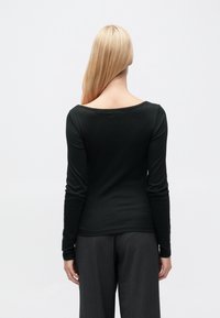 Μακρυμάνικη μαύρη μπλούζα με στρογγυλεμένο ντεκολτέ, κατασκευασμένη από απαλό ύφασμα, εφαρμοστή σιλουέτα, λεία υφή, φορεμένη με σκούρο γκρι παντελόνι.