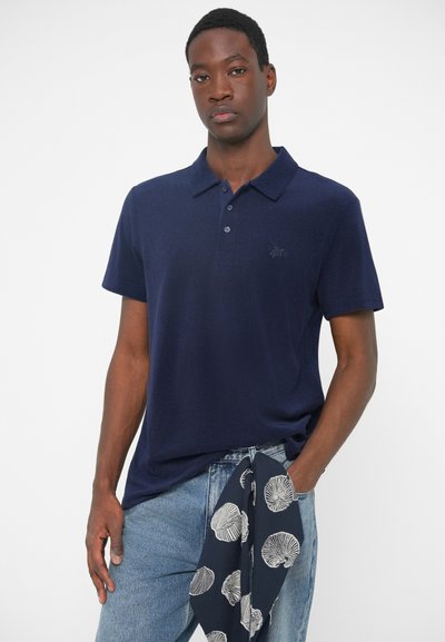 Tums zils polo krekls, īsas piedurknes, ar smalku logotipu uz krūšu. Valkāts ar gaiši ziliem džinsiem un rakstainu šalli, kas piesieta apkārt viduklim.