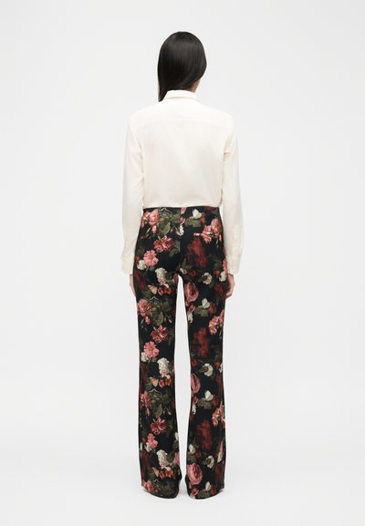 Nő háttal áll, fehér hosszú ujjú inget és fekete, virágmintás, bő szárú nadrágot visel egy egyszerű háttér előtt.