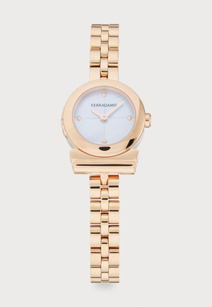 Gold-tone metal watch with a round face, pale blue dial, and minimalistic design. Features a linked bracelet and brand logo positioned on the face.