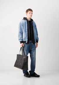 Chaqueta de mezclilla con sudadera negra, pantalones negros y zapatillas; llevando un gran bolso tote negro con el logotipo "HUGO" en relieve.