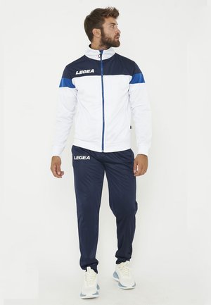 Homme portant une veste de sport blanche et bleu marine et un pantalon bleu marine assorti avec des baskets blanches, regardant sur le côté sur un fond blanc.