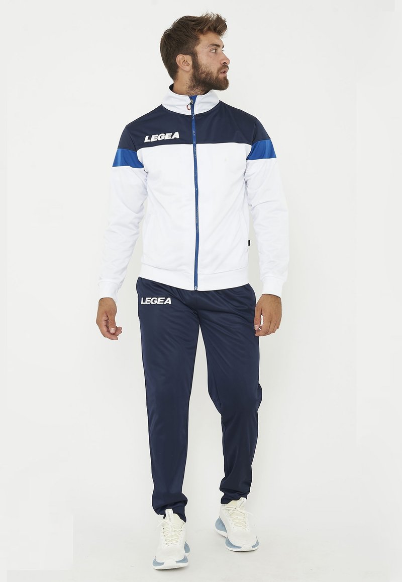Homme portant une veste de sport blanche et bleu marine et un pantalon bleu marine assorti avec des baskets blanches, regardant sur le côté sur un fond blanc.