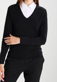 Schwarzer V-Ausschnitt-Pullover aus weichem Stoff, mit langen Ärmeln und tailliertem Design. Ein bemerkenswertes Detail ist ein kleines Logo auf der Brust.