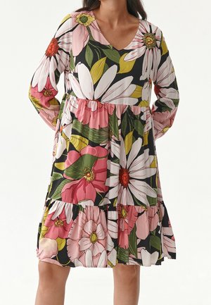 Femme portant une robe longue jusqu'aux genoux à manches longues avec un grand imprimé floral rose, blanc et jaune sur fond noir.