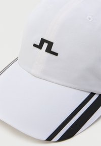 Valkoinen baseball-lippis, joka on valmistettu kankaasta, mustalla logolla ja kahdella mustalla raidalla lippalakin reunassa. Rakenne on jämäkkä ja pinta sileä.