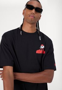 Μαύρη βαμβακερή μπλούζα κοντομάνικη, με κόκκινο γραφικό λογότυπο και καρτούν στο αριστερό στήθος. Λείος υφασμάτινος τύπος, χαλαρή εφαρμογή.