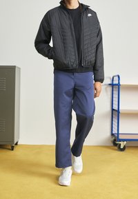 Svart vadderad jacka med dragkedja fram, marinblå byxor och vita sneakers. Har en diskret logotyp på bröstet och en upprättstående krage.