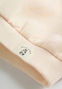 Puño de sudadera de color rosa claro, confeccionado en un tejido suave, que presenta una textura acanalada y una pequeña etiqueta tejida con un gráfico de una vaca y el texto "Milk Club".