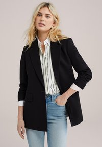 Černý tailored blazer s jedním knoflíkem, se strukturovanými rameny a mírným leskem, nasazený přes bílou košili s pruhy.