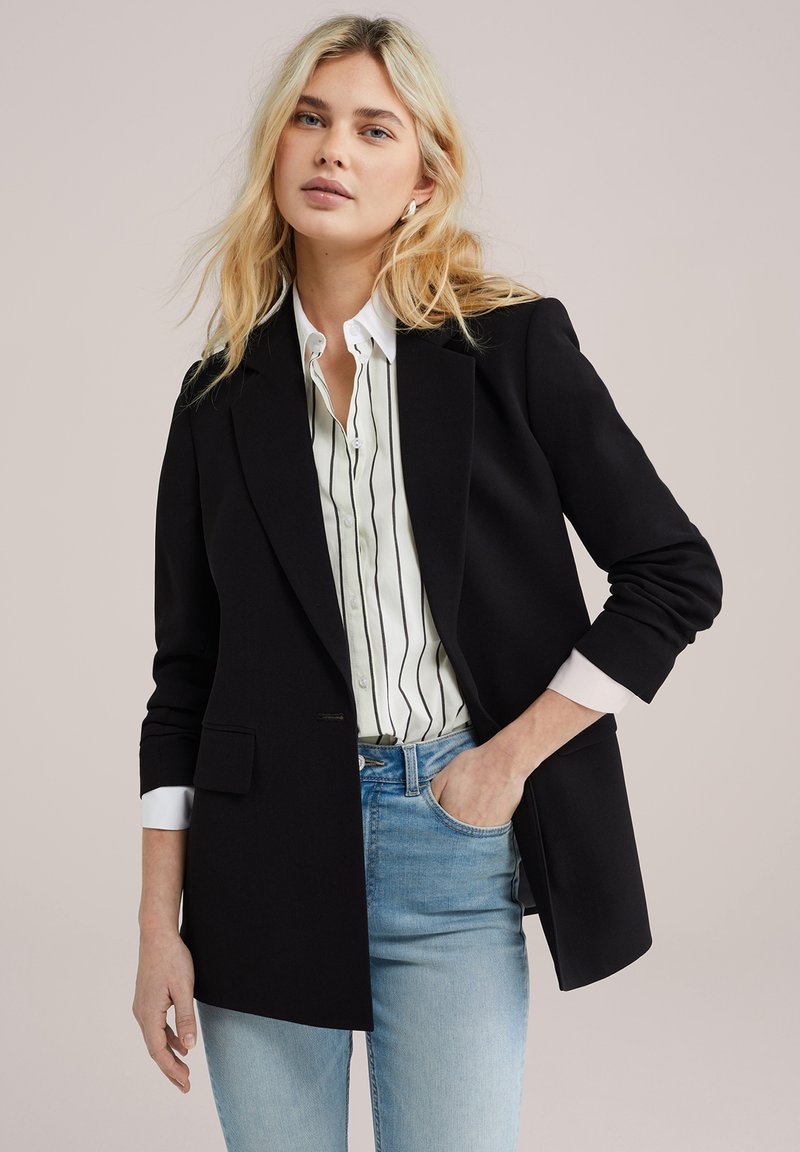 Černý tailored blazer s jedním knoflíkem, se strukturovanými rameny a mírným leskem, nasazený přes bílou košili s pruhy.