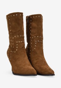 Botas de tobillo de ante marrón con punta afilada, que presentan tachuelas plateadas a lo largo de los lados y un cierre de cremallera en el tobillo interior.