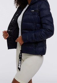 Granatowa kurtka puchowa z kapturem, zapinana na suwak, z prążkowanymi mankietami i akcentami z logo. Noszona na białej koszuli i jasnych prążkowanych szortach.