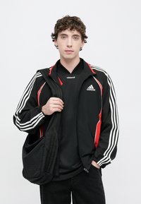 Čierna atletická bunda s bielymi a červenými akcentmi, s pásikavými rukávmi a predným zipsom. Nosená cez čierne tričko, čierna taška po boku.