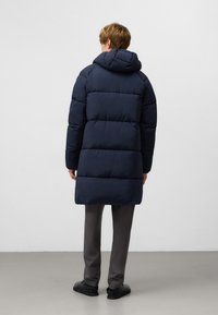 Granatowa kurtka puchowa z kapturem, z pikowanymi sekcjami i lekko błyszczącym wykończeniem. Nosi ją osoba w szarych spodniach i czarnych butach.