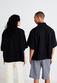 Sorte, oversized kortærmede skjorter har en afslappet pasform og krave, båret med cremefarvede bukser og blåstribede shorts, set fra bagenden.