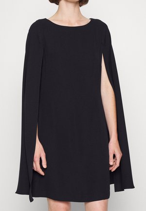 Woman wearing a knee-length black dress with split cape sleeves, standing against a plain light background.