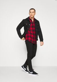 Schwarze Jeansjacke über einem rot-schwarzen Karohemd, kombiniert mit schwarzen Slim-Fit-Hosen und schwarzen Sneakers mit weißen Akzenten.