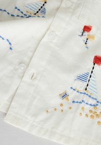 Chemise en tissu blanc ornée de voiliers brodés bleus et rouges et d'accents beige, avec des boutons visibles et des détails de couture texturés.