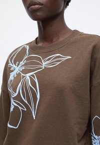 Gros plan d'une personne portant un sweat-shirt marron avec un grand motif floral bleu clair sur la poitrine et la manche.