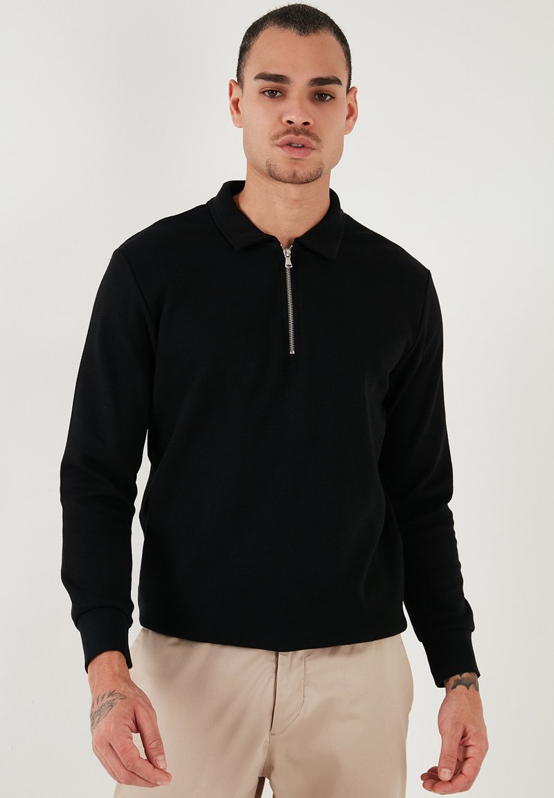 Camisa polo negra de manga larga con cremallera en el cuello, hecha de tela texturizada, con puños acanalados y un ajuste ceñido.