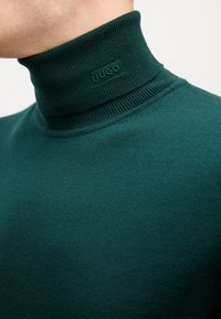 Dunkelgrüner Rollkragenpullover aus gerippter Strickware mit einem dezent erhabenen "HUGO"-Logo am Kragen. Glatte Textur.