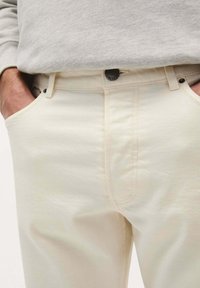 Pantaloni din bumbac bej deschis, cu o textură netedă, buzunare frontale și închidere cu buton metalic. Talie cu bucle pentru curea.