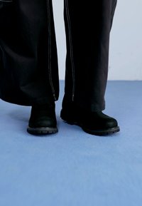 Bottines noires avec semelles en caoutchouc, texture lisse, bout rond, portées avec un pantalon noir à couture blanche, sur un fond bleu.
