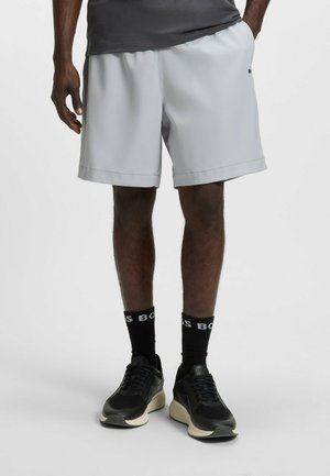 Lichtgrijze sportshorts met een gladde textuur en een elastische tailleband, gecombineerd met zwarte sneakers en zwarte sokken met witte tekst.