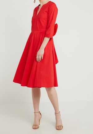 Red cotton dress with 3/4 sleeves, V-neckline, fitted bodice, and a flared skirt. Features a bow accent at the back. Worn with nude sandals.