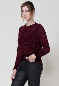 Burgunderfarbener Strickpullover mit strukturierter Oberfläche, gerippten Bündchen und Saum, im lässigen Schnitt und mit Rundhalsausschnitt, kombiniert mit schwarzen Hosen.