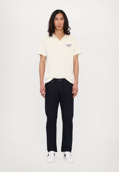 Polo shirt blanc cassé avec texte brodé, associé à un jean bleu foncé et des baskets blanches. Simple, coupe classique. Pas de motifs ni d'ornements.